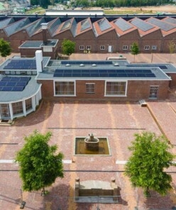 Luchtfoto van een bakstenen kantoorpand met zonnepanelen aan een plein met fontein.