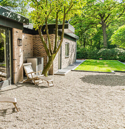 Moderne bakstenen bungalow met grindterras, houten ligstoel en een zonnig gazon in bosrijke omgeving.
